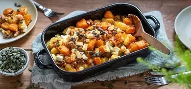 Kürbisgratin mit Kürbiswürfeln, Feta, Kürbiskernen und Gnocchi in Auflaufform auf Holztisch