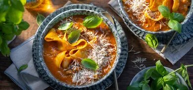 Kürbissuppe mit breiten Nudeln, geriebenem Parmesan und Basilikum in Schalen auf Holztisch