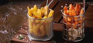 Zwei Metallkörbe mit Kartoffel- Pommes und Süßkartoffel-Pommes, serviert auf einem Holzbrett