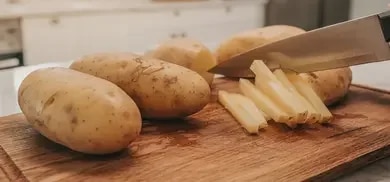 Rohe Kartoffeln liegen nebeneinander auf einem Holzbrett, eine davon wird gerade in Stifte geschnitten.]