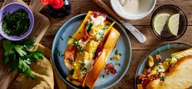 Sandwich mit Ruehrei, Speck und Kaese im Broetchen, daneben Kraeuter, Limettenscheiben und Sauce in Schuesseln