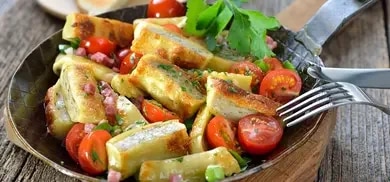 Maultaschen angerichtet in einer Pfanne auf einem Holzbrett mit Tomaten, Speck und Kräutern garniert mit Frühlingszwiebeln.