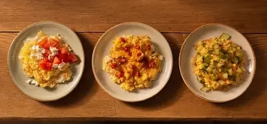 Drei kleine Teller mit verschiedenen Rührei-Variationen: eine mit Tomate/Feta, eine mit Chili/Maggi Würze Hot und eine mit Gemüse