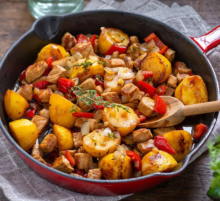 Kartoffelpfanne mit Fleischwürfeln, Paprika und Zwiebeln in einer Pfanne auf Holztisch
