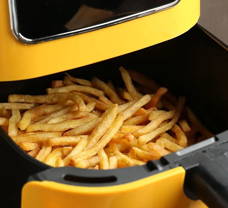 Eine moderne Heißluftfritteuse in einer hellen, freundlichen Küche. Aus dem geöffneten Korb quellen goldbraune Pommes