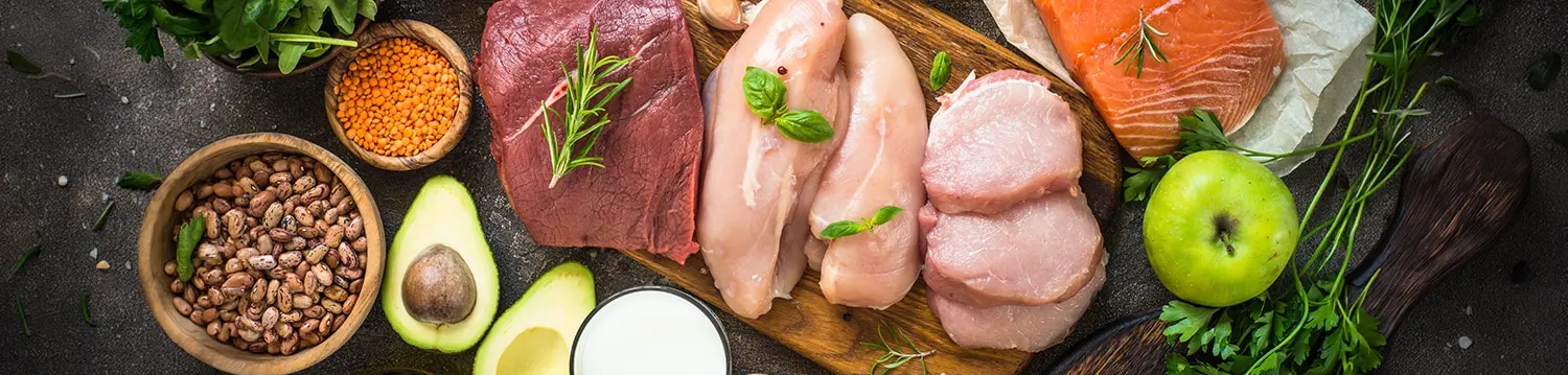 Auf einer dunklen Oberfläche liegen verschiedene proteinreiche Lebensmittel dekorativ angeordnet, darunter roher Lachs, Hähnc