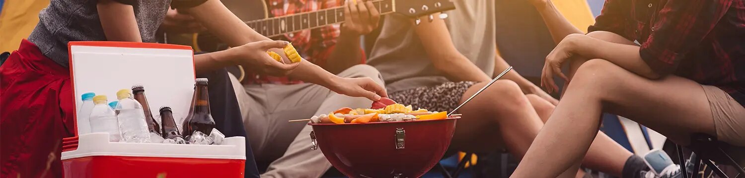Gruppe sitzt vor Zelt beim Grillen, runder Grill mit Essen, Kühlbox mit Flaschen und Person mit Gitarre
