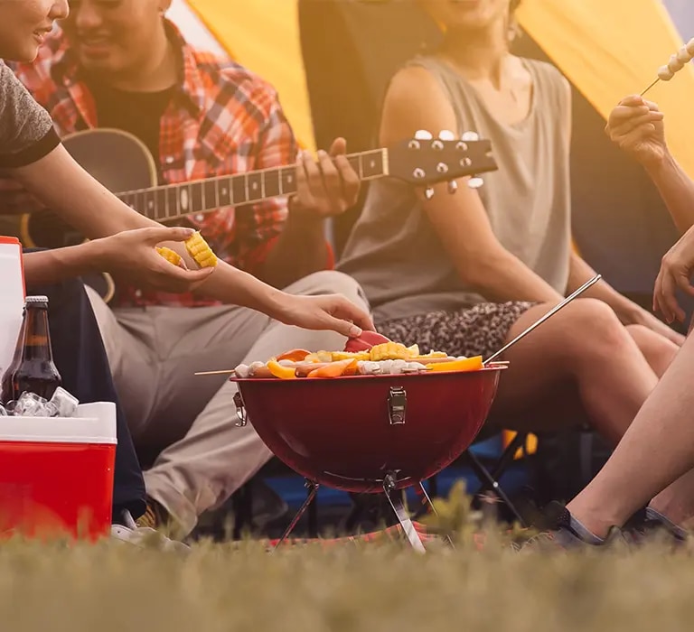 Gruppe sitzt vor Zelt beim Grillen, runder Grill mit Essen, Kühlbox mit Flaschen und Person mit Gitarre
