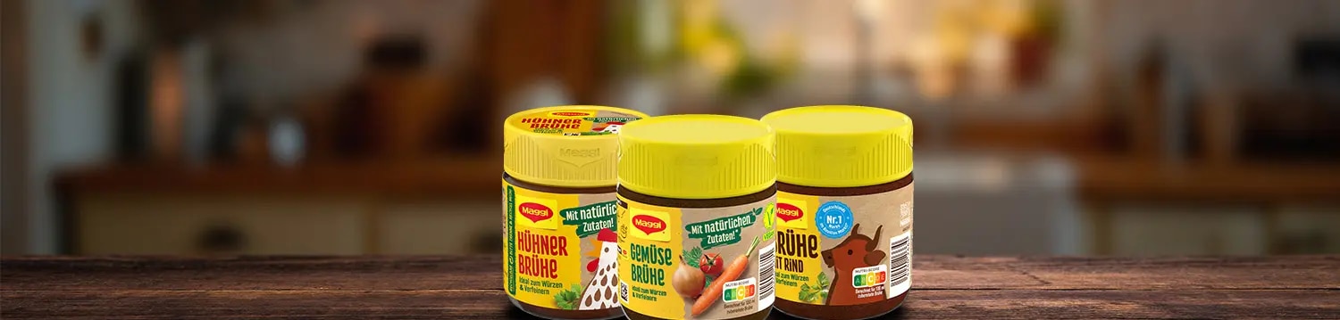 Drei Maggi Bruehe-Glaeser fuer Huehner-, Rinder- und Gemuese-Bruehe auf Holztisch mit Kuechenhintergrund