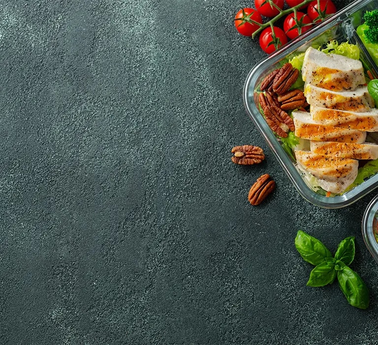 Zwei Meal-Prep-Boxen mit gegrilltem Haehnchen, Brokkoli, halbierten Eiern, Tomaten und Nuessen auf grauem Hintergrund