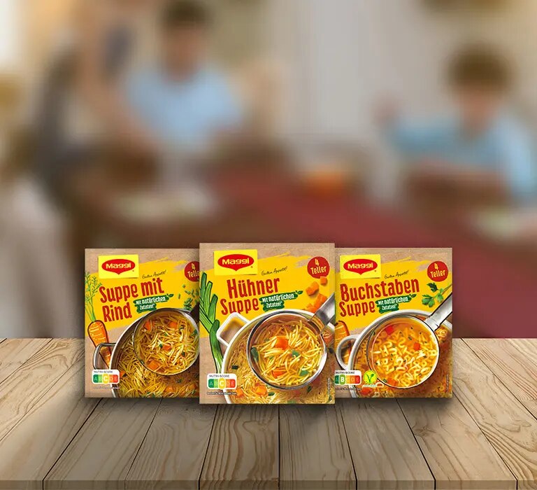 Drei Maggi Suppenverpackungen mit Rind-, Huehner- und Buchstabensuppe auf Holztisch vor Küchenhintergrund