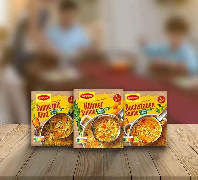 Drei Maggi Suppenverpackungen mit Rind-, Huehner- und Buchstabensuppe auf Holztisch vor Küchenhintergrund