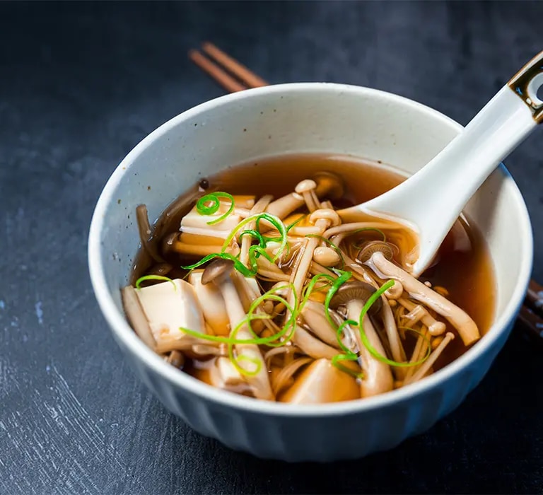 Schale mit Dashi-Brühe, Pilzen, Tofu und Frühlingszwiebeln, daneben Löffel und Essstäbchen