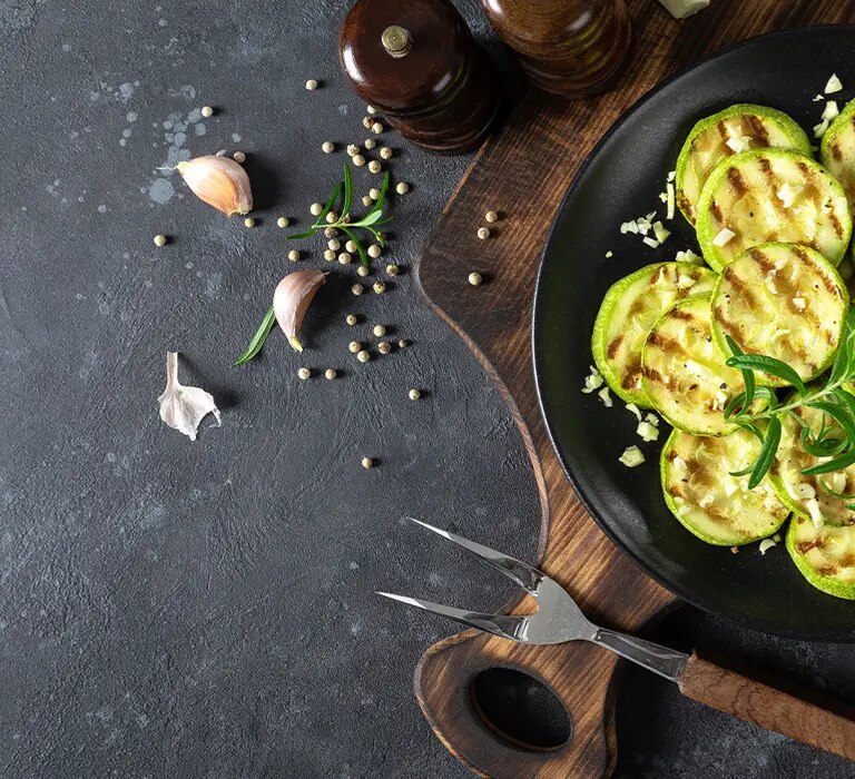 Gegrillte Zucchinischeiben auf schwarzem Teller mit Rosmarin, Knoblauch und Gewürzen auf Holzbretterfläche