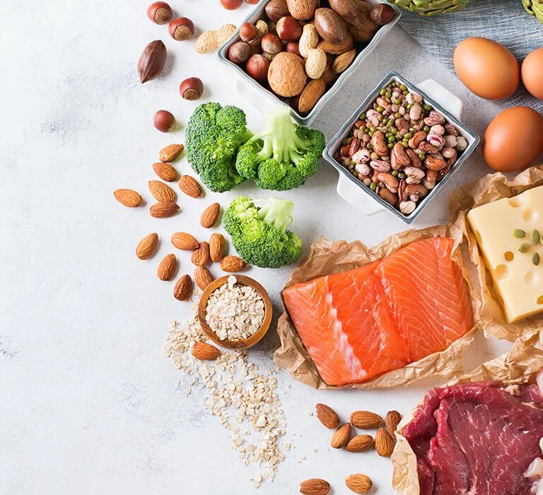 Lachs, Fleisch, Eier, Bohnen, Nüsse, Brokkoli, Käse und Joghurt auf heller Oberfläche.