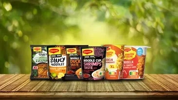Maggi Instant-Nudeln Sortiment mit Bechern und Päckchen auf Holztisch vor unscharfem grünem Hintergrund