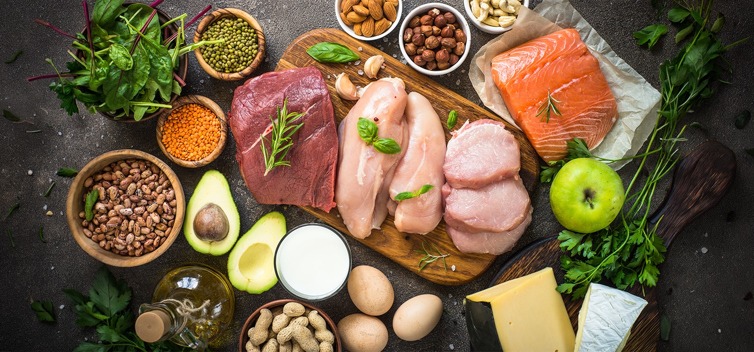 Auf einer dunklen Oberfläche liegen verschiedene proteinreiche Lebensmittel dekorativ angeordnet, darunter roher Lachs, Hähnc
