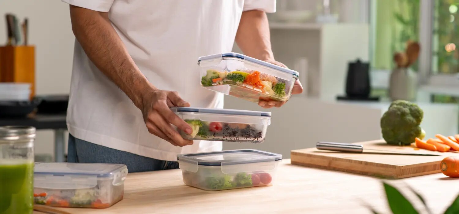 Person stapelt Meal-Prep-Boxen mit Gemuese auf Kuechentisch neben Schneidebrett mit Brokkoli und Karotten