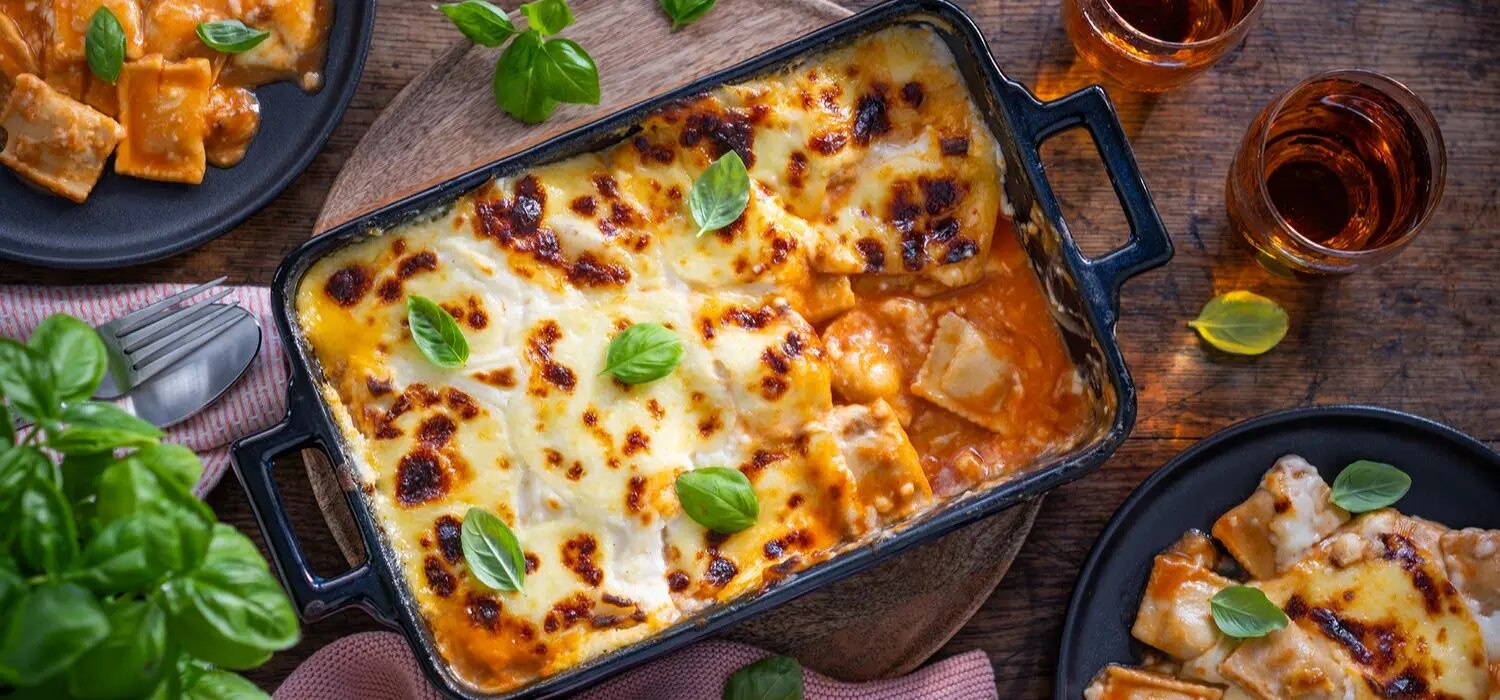 Ravioli-Lasagne in Auflaufform mit geschmolzenem Kaese, Tomatensauce und Basilikum auf Holztisch