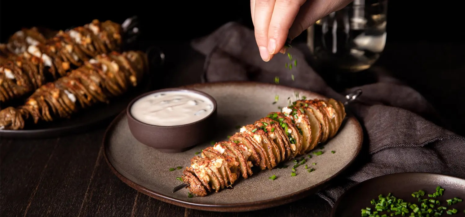 Kartoffelspiralen am Spieß mit Feta und Kräutern auf Teller, daneben Schale mit Dip, Hand streut Kräuter