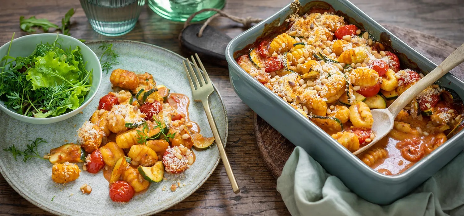 Gnocchi-Gratin mit Tomatensauce in Auflaufform und auf Teller mit Salat auf Holztisch