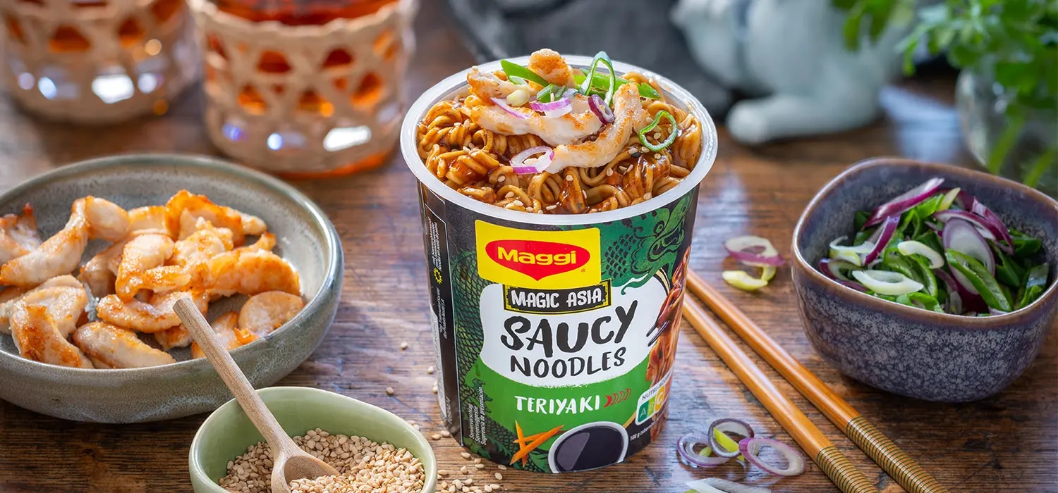 MAGGI Saucy Noodles Teriyaki Cup mit Haehnchenstreifen und Fruehlingszwiebeln auf Tisch mit Zutaten