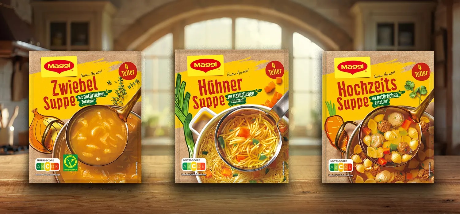 Drei Maggi Suppenpackungen mit Zwiebel-, Huehner- und Hochzeitssuppe auf Holztisch vor Kuechenhintergrund