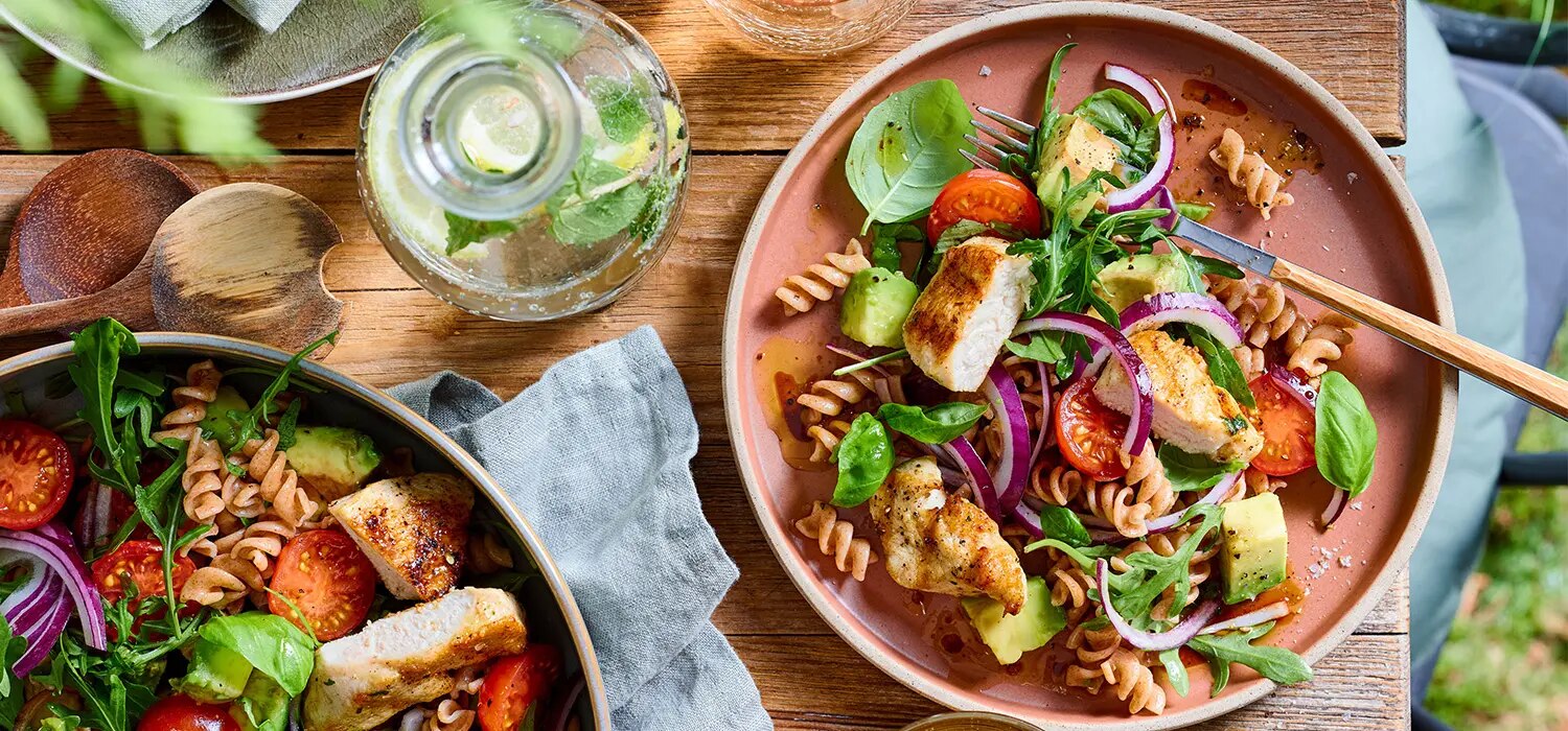 Nudelsalat mit gegrilltem Haehnchen, Avocado, Tomaten und roten Zwiebeln auf zwei Tellern