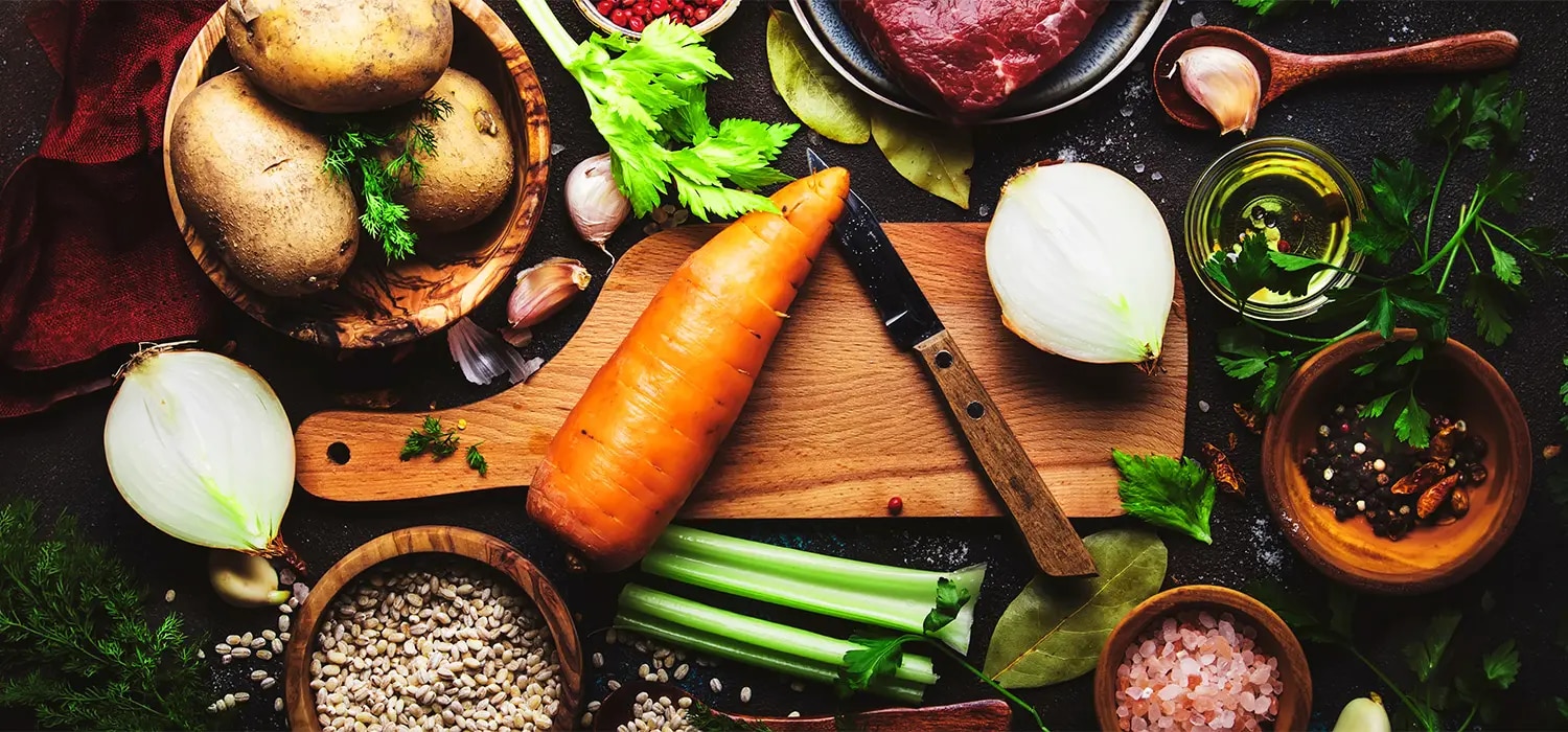 Karotte, Kartoffeln, Zwiebel, Sellerie, Rindfleisch, Knoblauch, Kraeuter und Gewuerze auf Schneidebrett in Draufsicht