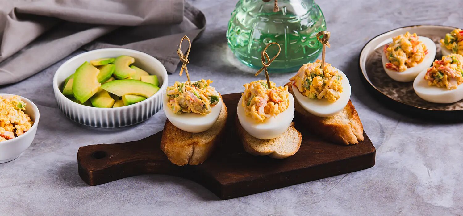 Gefüllte Eierhälften auf Baguettescheiben mit Spießen, Avocadoscheiben und Schüsseln auf Tisch angerichtet