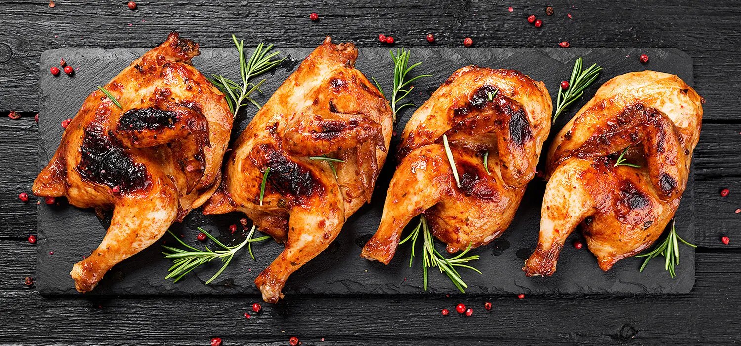 Vier gegrillte Hähnchenschenkel mit Grillspuren und Kräutern auf dunkler Platte angerichtet