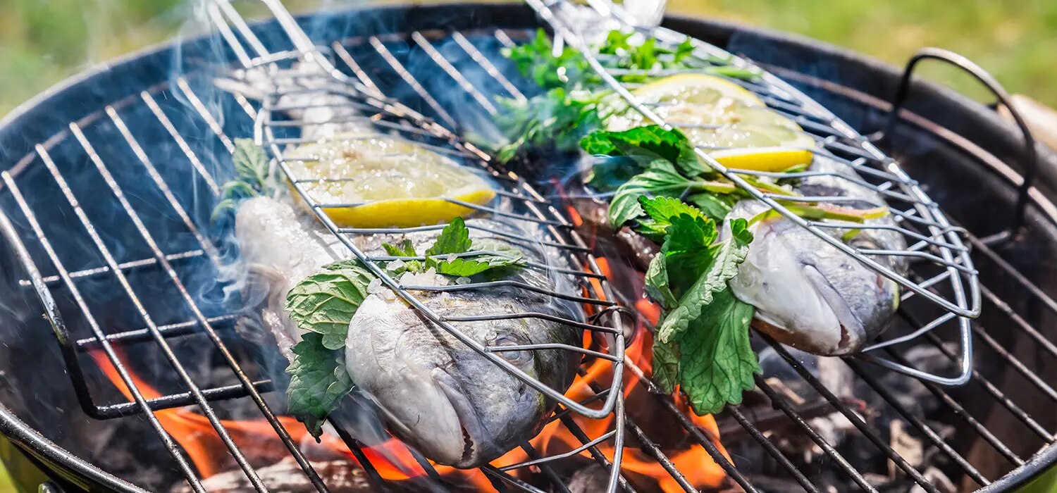 Fisch im Grillkorb mit Zitronenscheiben und Kraeutern ueber Holzkohlegrill mit Flammen und Rauch