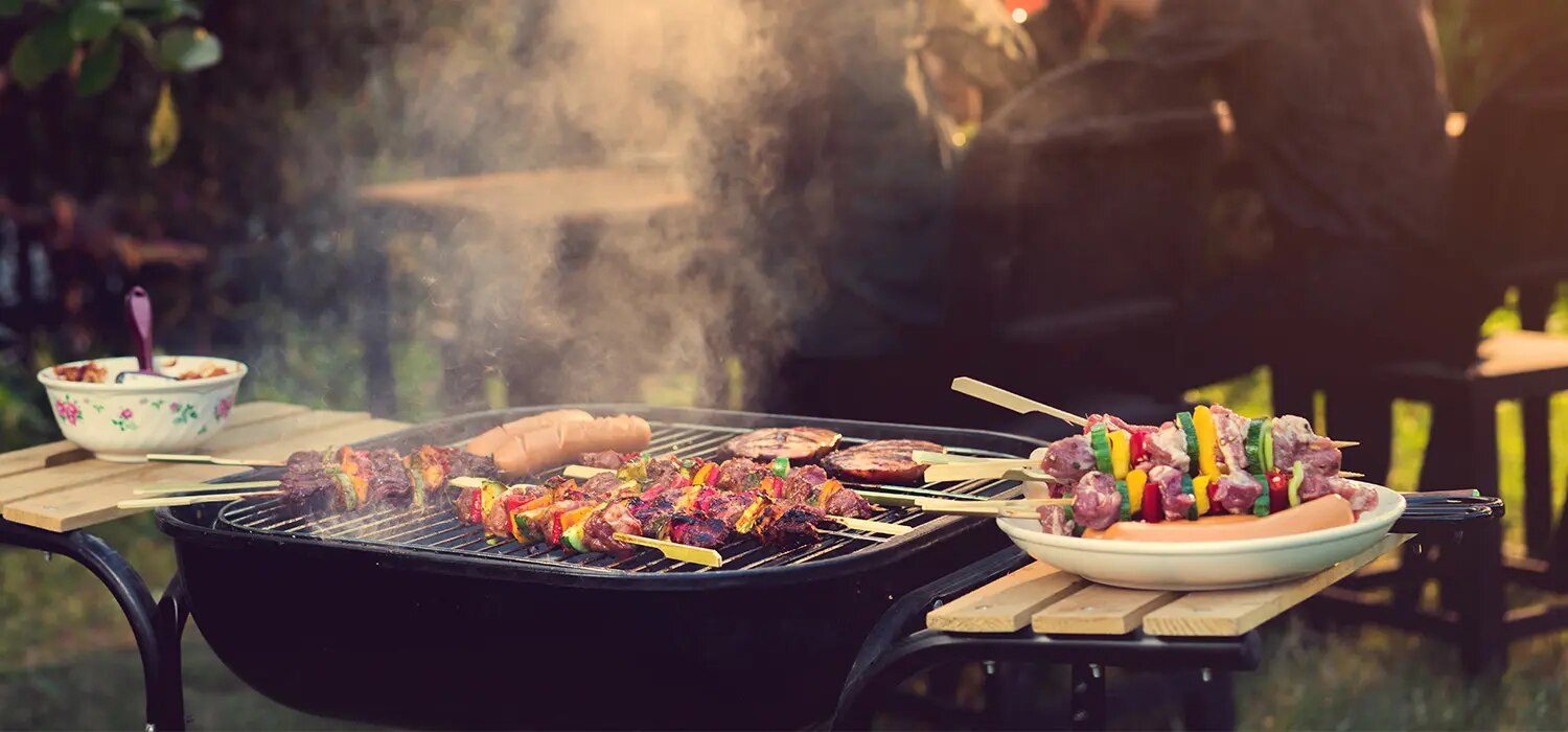 Grill mit Spießen aus Fleisch und Gemüse sowie Würstchen, Teller mit bunten Spießen daneben, Rauch über Grill