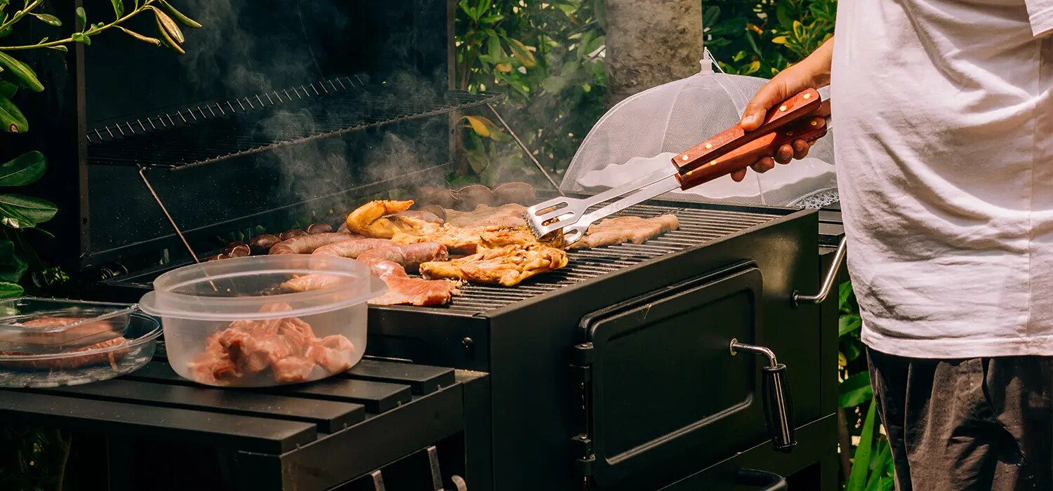 Person grillt Fleisch und Würstchen auf Holzkohlegrill, daneben Schalen mit rohem Fleisch und Grillzange