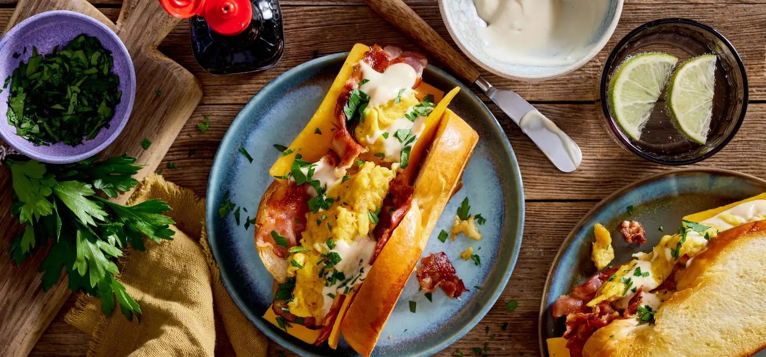 Sandwich mit Ruehrei, Speck und Kaese im Broetchen, daneben Kraeuter, Limettenscheiben und Sauce in Schuesseln