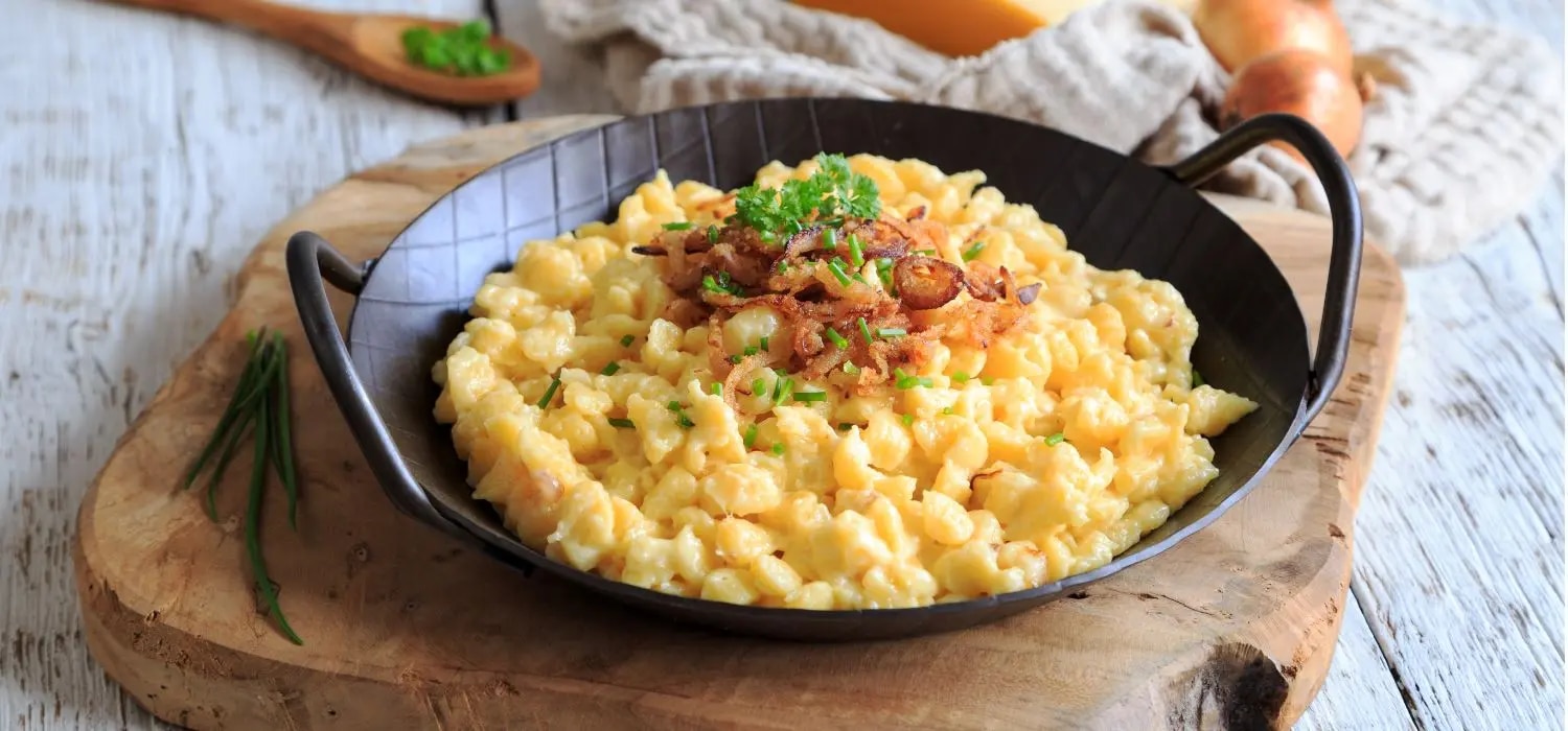 Käsespätzle mit Röstzwiebeln garniert mit Kräutern serviert in einer dunklen topfähnlichen Schüssel auf einem Holzbrett.