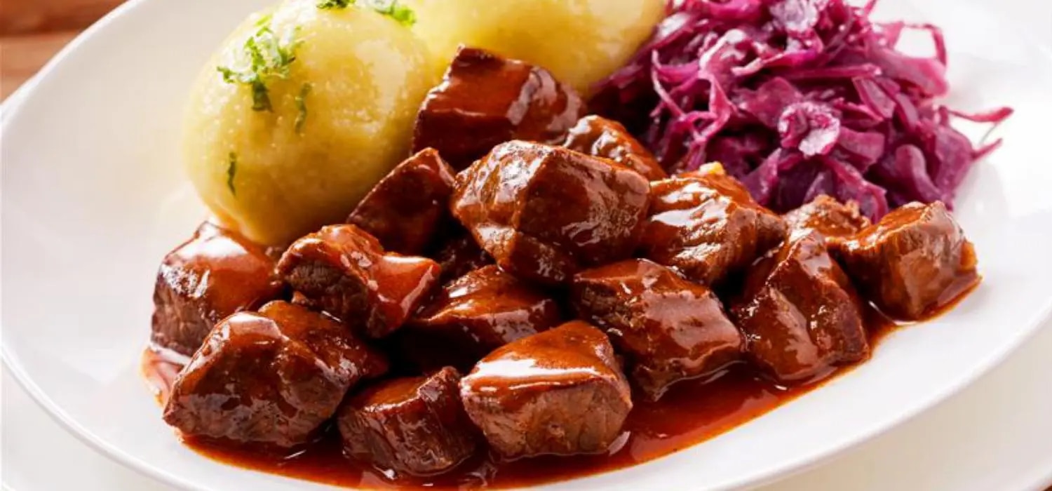 Rindergulasch mit Kartoffelknoedel und Rotkohl in Sauce in einer Schuessel serviert.