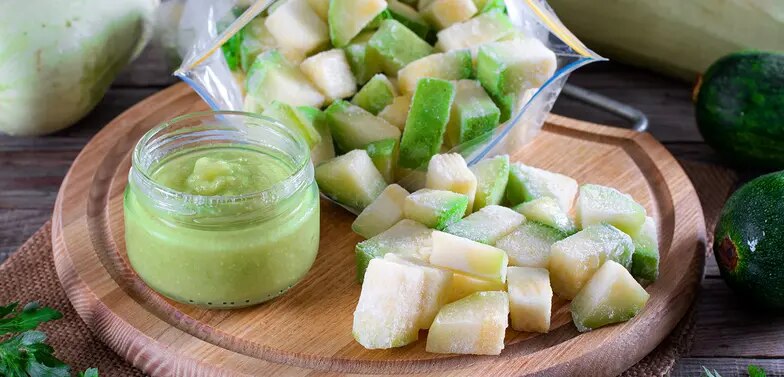Gefrorene Zucchiniwürfel in Beutel und Schale mit Zucchinipüree im Glas auf Holzbrett
