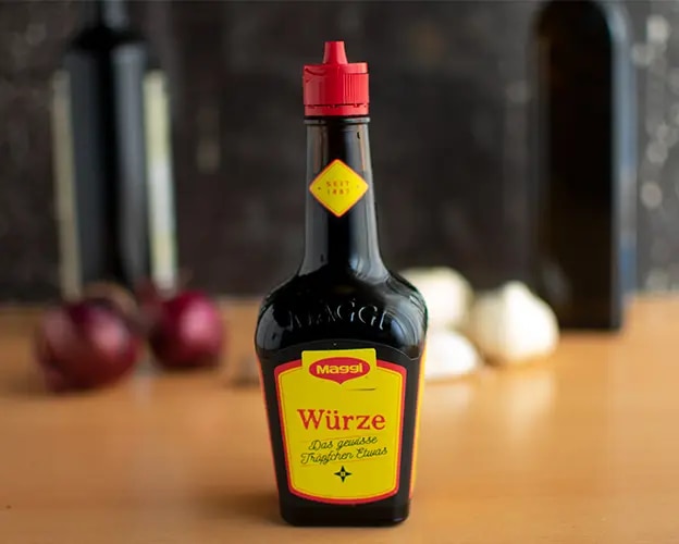 Flasche Maggi Würze auf einem Tisch, unscharfer Hintergrund mit Zwiebeln und Knoblauch.