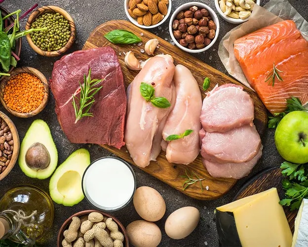 Auf einer dunklen Oberfläche liegen verschiedene proteinreiche Lebensmittel dekorativ angeordnet, darunter roher Lachs, Hähnc