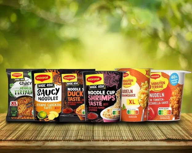 Maggi Instant-Nudeln Sortiment mit Bechern und Päckchen auf Holztisch vor unscharfem grünem Hintergrund