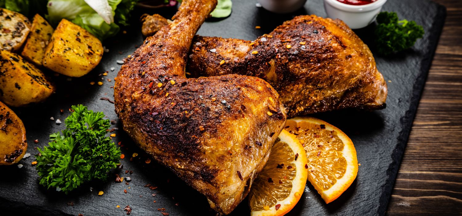 Zwei knusprig gebackene Hühnerschenkel mit Kartoffeln, Salat, Zitronenscheiben, Kräutern und Dips serviert auf einer Steinplatte.