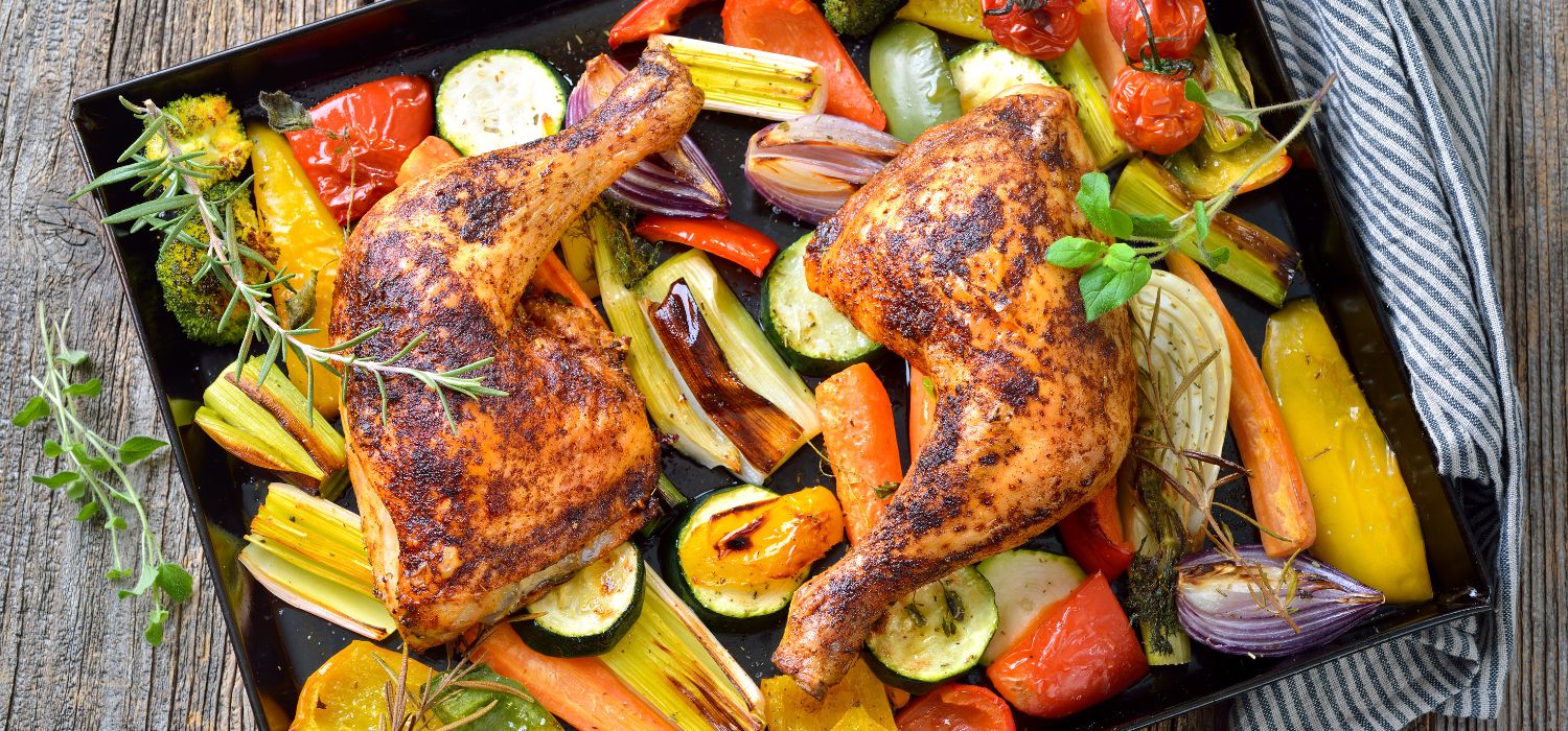 Zwei knusprig gebackene Hühnerkeulen mit mediterranem Gemüse heiß auf dem Backblech serviert.