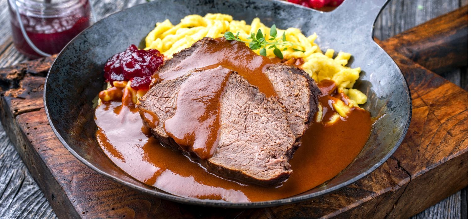 Geschmorter und marinierter Sauerbraten vom Rind mit Spätzle und Preiselbeermarmelade in Sauce serviert in einer rustikalen gusseisernen Bratpfanne.