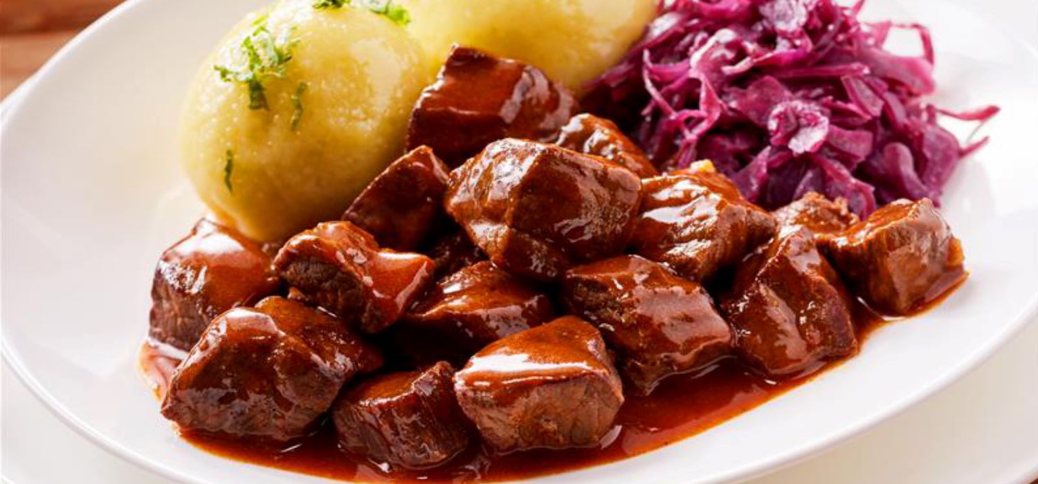 Rindergulasch mit Kartoffelknoedel und Rotkohl in Sauce in einer Schuessel serviert.